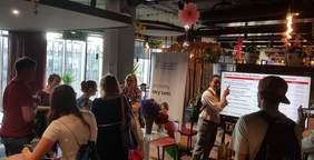 Pokaż zdjęcie: Kobieta przy stoisku Oddziału Wielkopolskiego PFRON udziela informacji grupa pięciu osób, które znajdują się w pobliżu. W tle widoczny jest baner PFRON, a po prawej stronie ekran z wyświetloną agendą Targów Pracy, którą analizują dwie osoby.