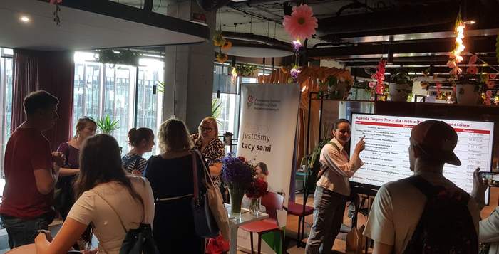 Pokaż zdjęcie: Kobieta przy stoisku Oddziału Wielkopolskiego PFRON udziela informacji grupa pięciu osób, które znajdują się w pobliżu. W tle widoczny jest baner PFRON, a po prawej stronie ekran z wyświetloną agendą Targów Pracy, którą analizują dwie osoby.