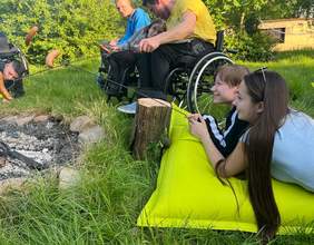 Pokaż zdjęcie: Sześć osób poruszających się na wózku znajduje się na trawniku, przy ognisku i trzyma w dłoniach metalowe kijki, a na nich kiełbaski. 