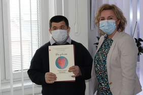 Pokaż zdjęcie: Mężczyzna i kobieta stoją na tle okna. Mężczyzna  trzyma dyplom z sercem. Obok niego stoi kobieta.