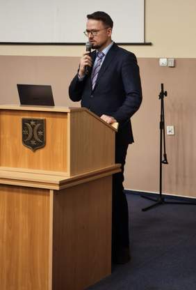 Pokaż zdjęcie: Mężczyzna w garniturze i krawacie stojący za drewnianą mównicą, przemawiający do mikrofonu. Na mównicy znajduje się herb ze złotymi gwiazdami na czarnym tle. Za nim widać ekran i laptop.