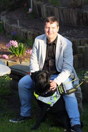 Pokaż zdjęcie: Mężczyzna siedzący na ławce, dotyka siedzącego przy nim psa przewodnika.