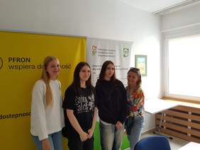 Pokaż zdjęcie: 3 uczennice oraz nauczycielka stoją na tle banerów PFRON w siedzibie Oddziału PFRON w Zielonej Górze