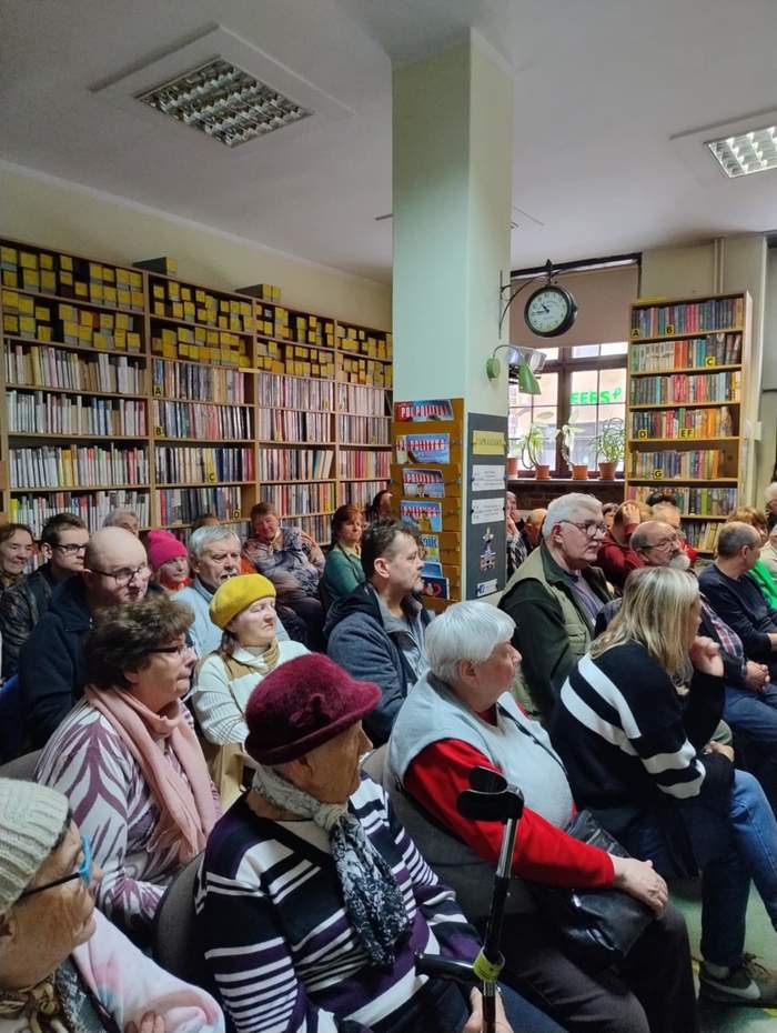 Uczestnicy spotkania siedzą na krzesłach. Za nimi widać regały z książkami.