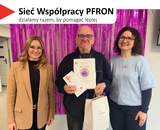 Na górze, na białym tle, widoczny jest czerwony, trójkątny element. Obok znajduje się tekst: 'Sieć Współpracy PFRON', a poniżej napis: 'działamy razem, by pomagać lepiej'. W centrum stoi mężczyzna trzymający dyplom oraz białą torbę z logo PFRON. Po lewej towarzyszy mu kobieta o długich blond włosach w okularach. Po prawej stoi kobieta z ciemnymi, kręconymi włosami w okularach, ubrana w jasnoniebieski sweter.