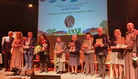 Pokaż zdjęcie: Wręczenie medali św. Brata Alberta. Na scenie laureaci, organizatorzy oraz prowadzący uroczystość. W tle ekran z napisem "Albertiana"