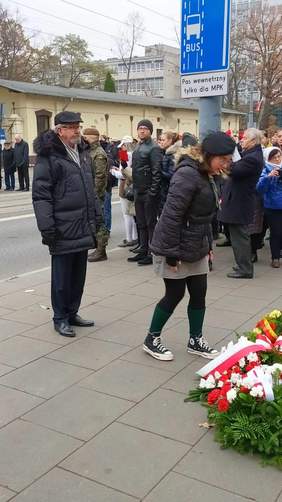 Pokaż zdjęcie: Po prawej stronie leżą biało-czerwone kwiaty. Przed nimi pochyla sie harcerka, a za nią stoi mężczyna, który jest dyrektorem Łódzkiego Oddziału PFRON. W tle widać tłum ludzi.  