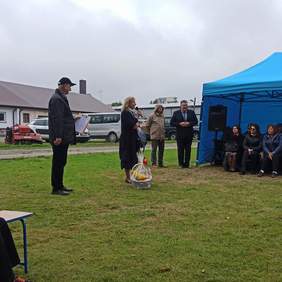 Pokaż zdjęcie: Zdjęcie na zewnątrz, przy niebieskim namiocie pod którym siedzą ludzie, na trawie stoją cztery osoby