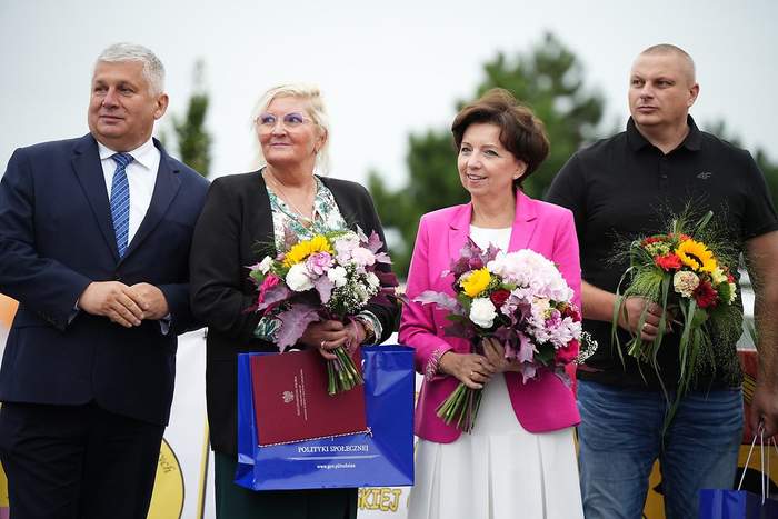 Pokaż zdjęcie: Na scenie, na tle drzew liściastych stoją dwie kobiety wraz z dwójką mężczyzn. Obie kobiety, jak i mężczyzna po prawej stronie trzymają w dłoniach bukiety kwiatów. Kobieta stojąca po lewej stronie trzyma w dłoniach także teczkę z godłem Polski i napisem: „Rzeczpospolita Polska” oraz granatową, papierową torbę wykorzystywaną na potrzeby reprezentacyjne Ministerstwa Rodziny i Polityki Społecznej.