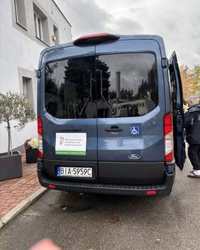 Pokaż zdjęcie: Niebieski mikrobus marki Ford Transit, przystosowany do przewozu osób z niepełnosprawnościami. Na drzwiach naklejka z logo Państwowego Funduszu Rehabilitacji Osób Niepełnosprawnych (PFRON) oraz informacją o dofinansowaniu zakupu ze środków Funduszu. Na drzwiach widoczny jest również symbol wózka inwalidzkiego.