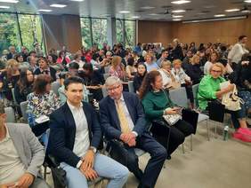 Pokaż zdjęcie: W sali w rzędach siedzą na krzesłach ludzie. Jest ich kilkadzisiąt. W tle widać okna, a za ich szybami bujną roślinność. 