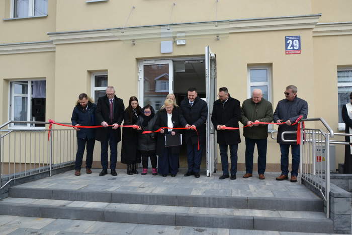 Pokaż zdjęcie: Grupa osób stoi przed wejściem do budynku i wspólnie przecina czerwoną wstęgę podczas uroczystego otwarcia. Do wejścia prowadzą schody i podjazd z poręczą.