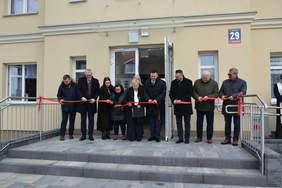 Pokaż zdjęcie: Grupa osób stoi przed wejściem do budynku i wspólnie przecina czerwoną wstęgę podczas uroczystego otwarcia. Do wejścia prowadzą schody i podjazd z poręczą.
