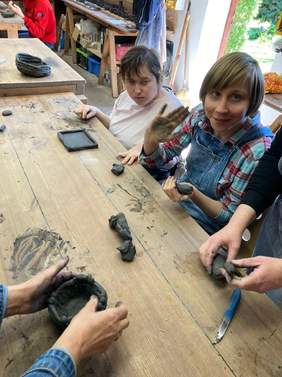 Pokaż zdjęcie: kilka osób siedzi przy dużym drewnianym stole na którym jest  rozmieszczona glina do wyrobów prac plastycznych przez uczestników warsztatów