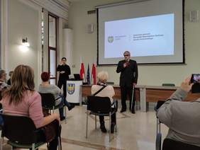 Pokaż zdjęcie: Na dużej sali wysoki mężczyzna ubrany w czarny garnitur w jednej ręce trzyma mikrofon w drugiej białą laskę obok stoi kobieta ubrana na czarno i tłumaczy na język migowy przed nimi siedzą ludzie w rzędach