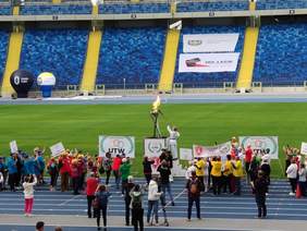Pokaż zdjęcie: Zawodnik ubrany na biało trzyma pochodnię i zapala znicz stojący na podwyższeniu przyglądają się temu zawodnicy i fotoreporterze robiąc zdjęcia