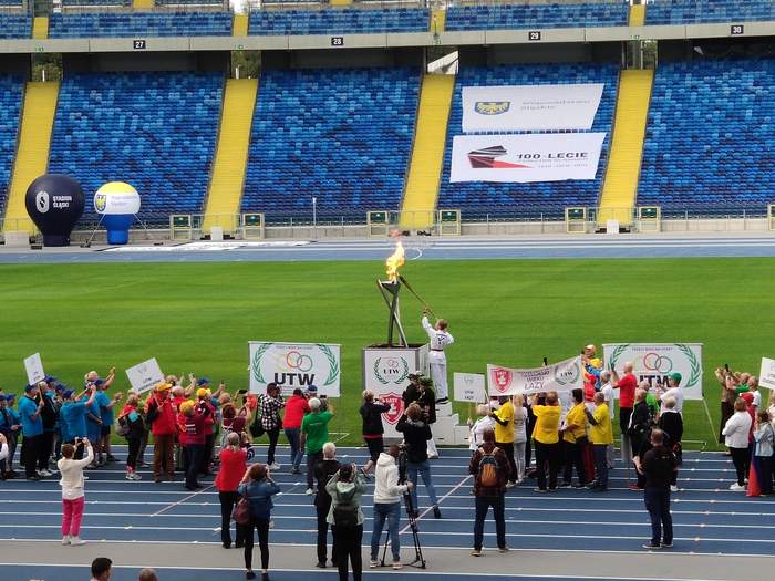 Pokaż zdjęcie: Zawodnik ubrany na biało trzyma pochodnię i zapala znicz stojący na podwyższeniu przyglądają się temu zawodnicy i fotoreporterze robiąc zdjęcia