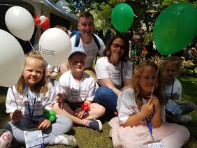 Grupa dzieci siedząca na trawie. Trzymają balony z logo PFRON. Wśród nich dorosła uśmiechnięta  kobieta. 