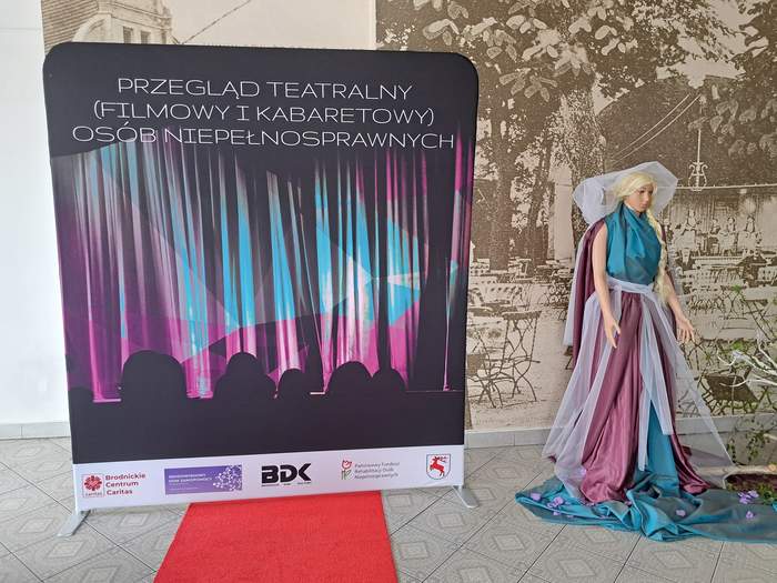 Baner reklamujący V Przegląd, a  obok manekin ubrany w kolorowy, teatralny strój