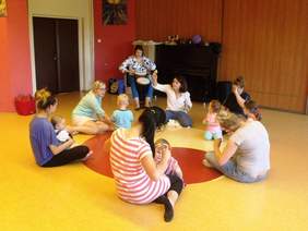 Pokaż zdjęcie: Grupa dzieci i dorosłych siedząca na podłodze, w tle na krześle siedzi pani grająca na bębnach