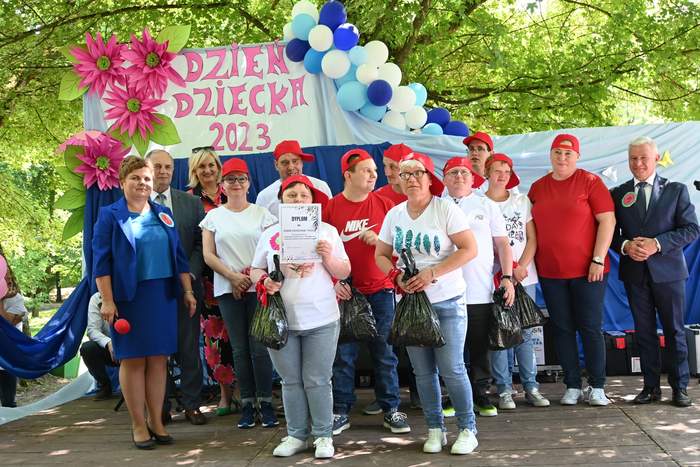 Pokaż zdjęcie: Na scenie stoją artyści ubrani w białe podkoszulki oraz jeansy. Na głowach mają czerwone basebolówki.  Pozują do pamiątkowego zdjęcia z z panią dyrektor Domu Pomocy Społecznej