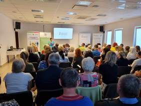 Pokaż zdjęcie: Zdjęcie podczas konferencji zrobione z tyłu sali, widać osoby biorące udział w konferencji oraz prelegenta. Na ścianie ekran z prezentacją.