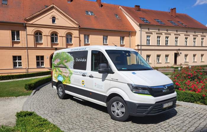 Pokaż zdjęcie: Na placu, przed dwukondygnacyjnym budynkiem ze skośnym dachem stoi biały bus z naklejką informującą, że pojazd przystosowany jest do przewozu osób z niepełnosprawnością. Na busie widnieje również naklejka z logo PFRON oraz z logo SOSW dla Dzieci Niewidomych w Owińskach. Ponadto pojazd oklejony został zarówno wizualizacją placówki oraz jej otoczenia, jak i następującymi sformułowaniami: „www.powiat.poznan.pl” oraz „powiat poznański”. Na placu przed ośrodkiem możemy dostrzec także kwitnące krzewy czerwonych róż, niski żywopłot z bukszpanu oraz jedną drewnianą ławkę.