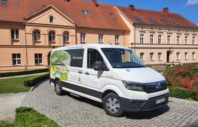 Pokaż zdjęcie: Na placu, przed dwukondygnacyjnym budynkiem ze skośnym dachem stoi biały bus z naklejką informującą, że pojazd przystosowany jest do przewozu osób z niepełnosprawnością. Na busie widnieje również naklejka z logo PFRON oraz z logo SOSW dla Dzieci Niewidomych w Owińskach. Ponadto pojazd oklejony został zarówno wizualizacją placówki oraz jej otoczenia, jak i następującymi sformułowaniami: „www.powiat.poznan.pl” oraz „powiat poznański”. Na placu przed ośrodkiem możemy dostrzec także kwitnące krzewy czerwonych róż, niski żywopłot z bukszpanu oraz jedną drewnianą ławkę.