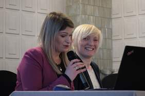 Pokaż zdjęcie: Dwie kobiety siedza przy stole konferencyjnym, jedna wpatruje się w laptopa, druga patrzy w obiektyw