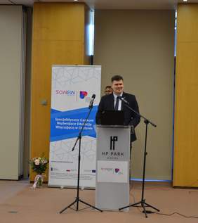 Pokaż zdjęcie: Mężczyzna stoi przed mównicą z dwoma mikrofonami. Z uśmiechem na twarzy wygłasza przemówienie