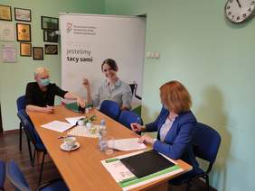 Pokaż zdjęcie: Dwie kobiety siedzą przy stole i rozmawiają, na stole są rozłożone dokumenty