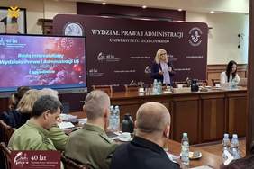 Pokaż zdjęcie: Kobieta w okularach i fioletowej marynarce przemawia do mikrofonu podczas spotkania Rady Interesariuszy na Wydziale Prawa i Administracji US. Stoi przed dużą bordową ścianką z logo uniwersytetu i napisem „40 lat”. Po jej lewej stronie znajduje się ekran multimedialny z napisem „Rada Interesariuszy Wydziału Prawa i Administracji US – I posiedzenie, 12 grudnia 2025 r.”. Na pierwszym planie widoczne są tyłem sylwetki słuchaczy, w tym osób w mundurach, siedzących przy konferencyjnym stole.