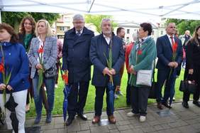 Pokaż zdjęcie: zdjęcie w plenerze osoby stoją pod zadaszeniem trzymają w ręku duży długi kwiat w kolorze czerwonym 