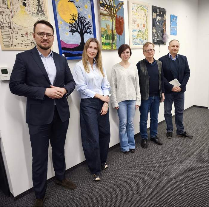 Pokaż zdjęcie: Pięć osób, trzech mężczyzn i dwie kobiety, stoi w szeregu, patrząc prosto na widza, wzdłuż ściany udekorowanej kolorowymi pracami artystycznymi, w tym tkaninami i obrazami.