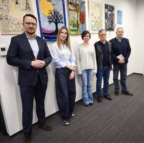 Pokaż zdjęcie: Pięć osób, trzech mężczyzn i dwie kobiety, stoi w szeregu, patrząc prosto na widza, wzdłuż ściany udekorowanej kolorowymi pracami artystycznymi, w tym tkaninami i obrazami.