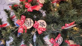 Pokaż zdjęcie: Zbliżenie na ozdoby choinkowe. Na choince wiszą drewniane bombki z ręcznie wypisanymi życzeniami, ozdobione czerwonymi kokardkami. W tle widoczne są igły choinki i delikatne światełka.