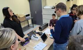 Pokaż zdjęcie: Prezentacja sprzętu dla osób z dysfunkcja wzroku. Na stole znajdują się lupy, maszyna do tyflografiki. Sprzęt dotykają uczestnicy spotkania.