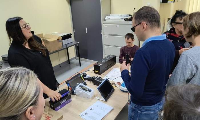 Pokaż zdjęcie: Prezentacja sprzętu dla osób z dysfunkcja wzroku. Na stole znajdują się lupy, maszyna do tyflografiki. Sprzęt dotykają uczestnicy spotkania.