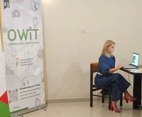 Pokaż zdjęcie: Kobieta siedzi przy stoliku z laptopem, po jej lewej stronie roll-up Ośrodka Wsparcia i Testów