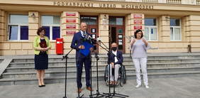 Cztery osoby, od lewej kobieta, mężczyzna mówiący do mikrofonów, mężczyzna na wózku inwalidzkim i kobieta tłumacząca na język migowy, za nimi budynek urzędu