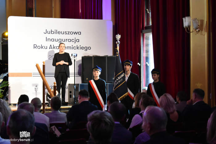 Na scenie widać tłumaczkę migową, za nią baner z napisem "Jubileuszowa Inauguracja Roku Akademickiego", po prawej stronie stoi poczet sztandarowy uczelni, tyłem do zdjęcia siedzą uczestnicy uroczystości