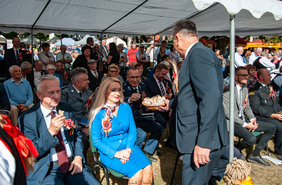 Pokaż zdjęcie: Zdjęcie gości zaproszonych na dożynki gminne, osoby te siedzą na krzesłach pod namiotem, mężczyzna w garniturze częstuje wszystkich kawałkami chleba z koszyka, który trzyma w rękach