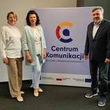 Dwie kobiety i jeden mężczyzna elegancko ubrani stoją przy rollupie z napisem i logo Centrum Komunikacji.