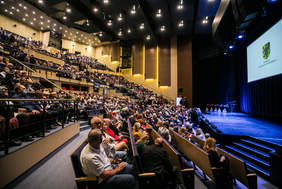 Pokaż zdjęcie: Widownia teatralna wypełniona gośćmi, po prawej stronie scena, nad którą wisi telebim z logo  Samorządu Województwa Pomorskiego. Na scenie leża elegancko opakowane Kosze wiklinowe - nagrody dla laureatów.