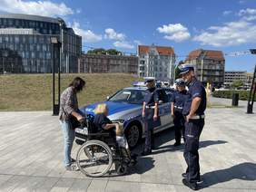 Pokaż zdjęcie: Na placu stoi radiowóz, przed nim stoi trzech umundurowanych policjantów. Kobieta siedząca na wózku inwalidzkim dotyka auta. Kobieta stojąca za nią trzymająca wózek mówi jej co dotyka w danym momencie.