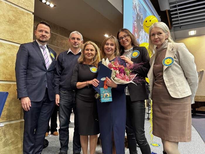 Pokaż zdjęcie: Grupa sześciu osób stoi blisko siebie na tle jasnej, fakturowanej ściany w auli lub sali konferencyjnej. W centralnym punkcie znajduje się kobieta w granatowej sukience, trzymająca bukiet różowych kwiatów oraz niebieski folder. Osoby mają przypięte do ubrań żółte przypinki z uśmiechniętym słońcem. Wszyscy patrzą w stronę obiektywu i uśmiechają się. W tle widoczne są żółte balony oraz fragment ekranu projekcyjnego.