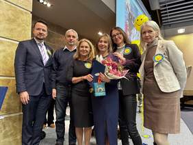 Pokaż zdjęcie: Grupa sześciu osób stoi blisko siebie na tle jasnej, fakturowanej ściany w auli lub sali konferencyjnej. W centralnym punkcie znajduje się kobieta w granatowej sukience, trzymająca bukiet różowych kwiatów oraz niebieski folder. Osoby mają przypięte do ubrań żółte przypinki z uśmiechniętym słońcem. Wszyscy patrzą w stronę obiektywu i uśmiechają się. W tle widoczne są żółte balony oraz fragment ekranu projekcyjnego.