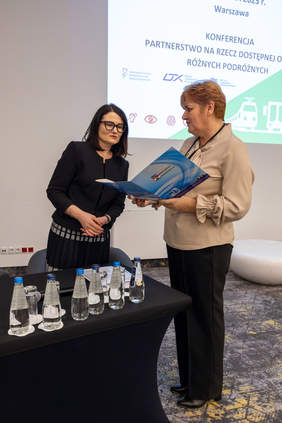Pokaż zdjęcie: Dwie kobiety. Od prawej p. Grażyna Lepianka przedstawicielka Międzygminnego Związku Komunikacyjnego przekazuje podpisaną deklarację p. Dorocie Habich Zastępcy Prezesa PFRON. W tle slajd z napisem Warszawa Konferencja Partnerstwo na rzecz dostępnej obsługi różnych podróżnych.