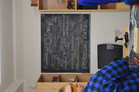 Pokaż zdjęcie: Na tablicy rozrysowany jest schemat pracy przy budowie karmiku dla ptaków. Są wymiary otworów w puszce, wymiary daszku oraz sposób wyznaczenia środka puszki do cyrkla żłobiarki. Są też wyznaczone osoby do poszczególnych czynności: Daszek – Ka