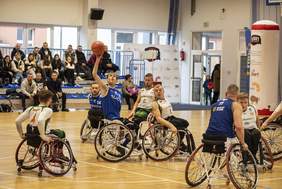 Pokaż zdjęcie: Mecz koszykówki na wózkach w hali sportowej. Zawodnik w niebieskiej koszulce podnosi piłkę do rzutu, otoczony przez graczy w biało-zielonych strojach i dwóch w niebieskich. W tle trybuny z widzami, banery promocyjne i drzwi wejściowe do hali.